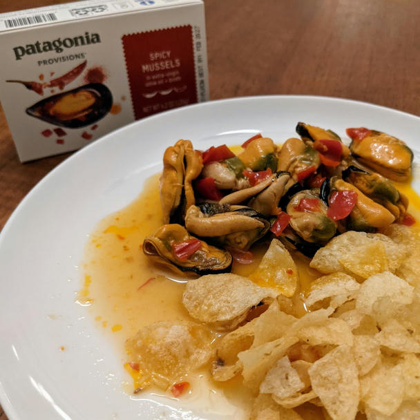 the empty box stands behind the finished plate on my teak veneer table. i turned the tin upside down, and the plate has a pool of broth in the center, mussels on one side, and chips on the other.

the mussels in their open tin, on a white ceramic plate. a bit of my black countertop in the corner, some potato chips at the top. a few mussels poke out of the broth; a mish mosh of chili pieces are mixed in. the broth has gorgeous droplets of red chili-infused oil in it
