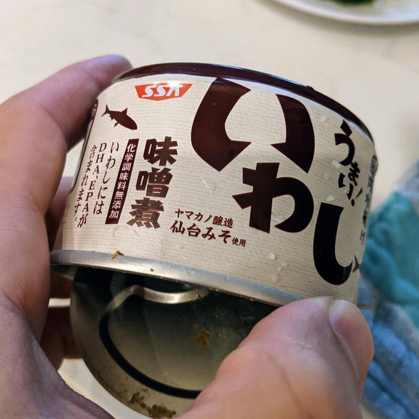 my fingers grasping the open beige can with brown writing saying 'iwashi' in hiragana, meaning sardines
