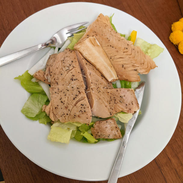 a white blate on a teak-veneer table. several chunks of salmon sit atop romaine lettuce, yellow bell pepper, and white onion. the salmon fillets have split at the lateral line and started to flake apart; some skin is folded up on one of the fillets. the pink flesh is interrupted by many specks of flavorful black pepper
