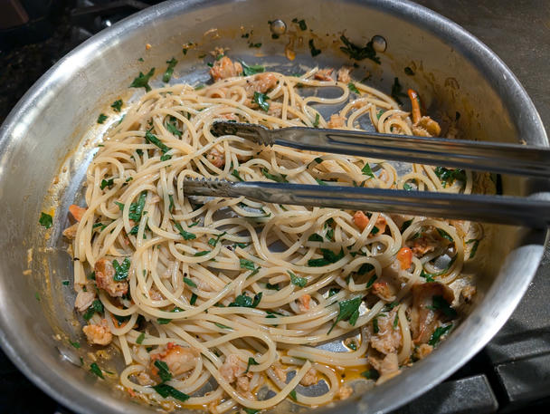 metal tongs rest on the side of broad saute pan, their serrated tips grabbing at a tangle of spaghetti. chunks of creamy red lobster and flecks of green parsley fill the shining, steel pan.
