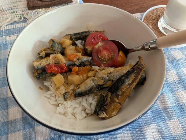 a white bowl with a blue outside holds short-grain white rice topped with five medium-small sardines, with red pepper and potato in the coconut curry sauce. a few tomato halves garnish. the whole ensemble is on a blue-and-white checkered placemat; a glass of kefir is on a coaster on the side.
